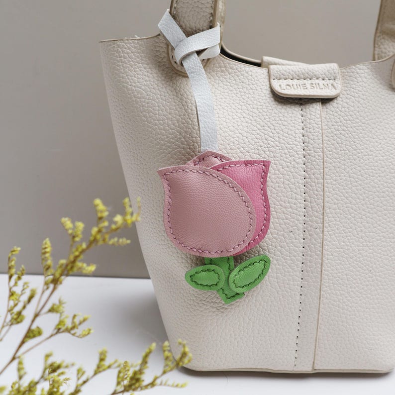 May include: A cream-colored leather handbag with a pink and light pink leather tulip-shaped charm attached. The charm has green leaves and a white leather strap. The handbag has the text "LOUIE SILMA" on the front.