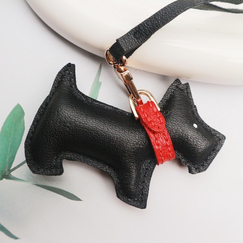 May include: A black leather dog-shaped keychain with a red collar and gold-colored hardware. The keychain has a black strap and is designed to resemble a Scottish Terrier. The dog's eye is a small white dot.