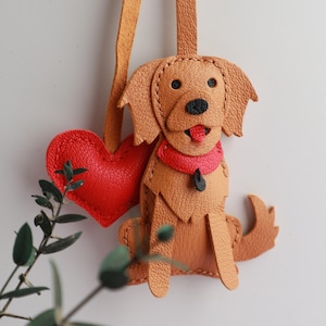 May include: A handcrafted leather dog charm with a red heart. The charm features a golden retriever design with a red collar and tongue. The charm is attached to a tan leather strap, perfect for a keychain or bag accessory.