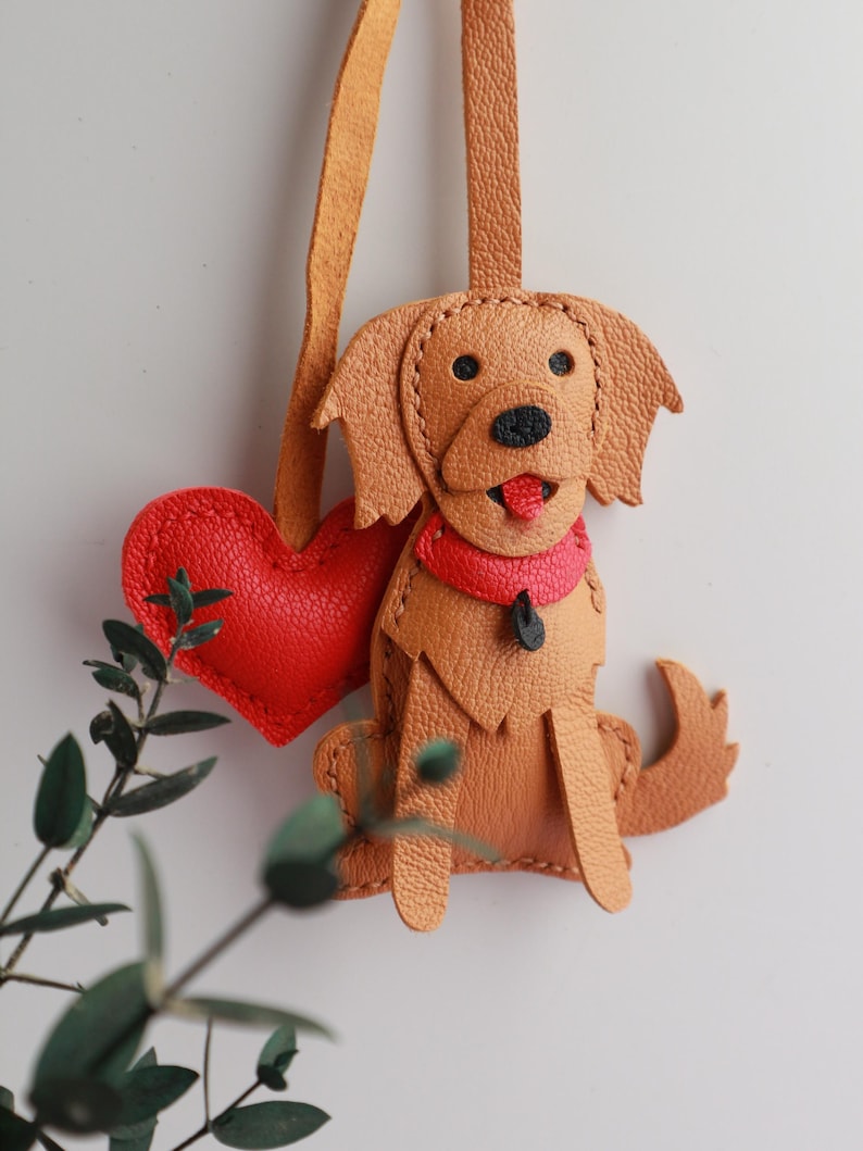 May include: A handcrafted leather dog charm with a red heart. The charm features a golden retriever design with a red collar and tongue. The charm is attached to a tan leather strap, perfect for a keychain or bag accessory.