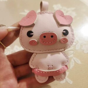 May include: A pink pig keychain made of leather with pink heart-shaped ears and a pink snout. The pig has black eyes and a stitched smile.