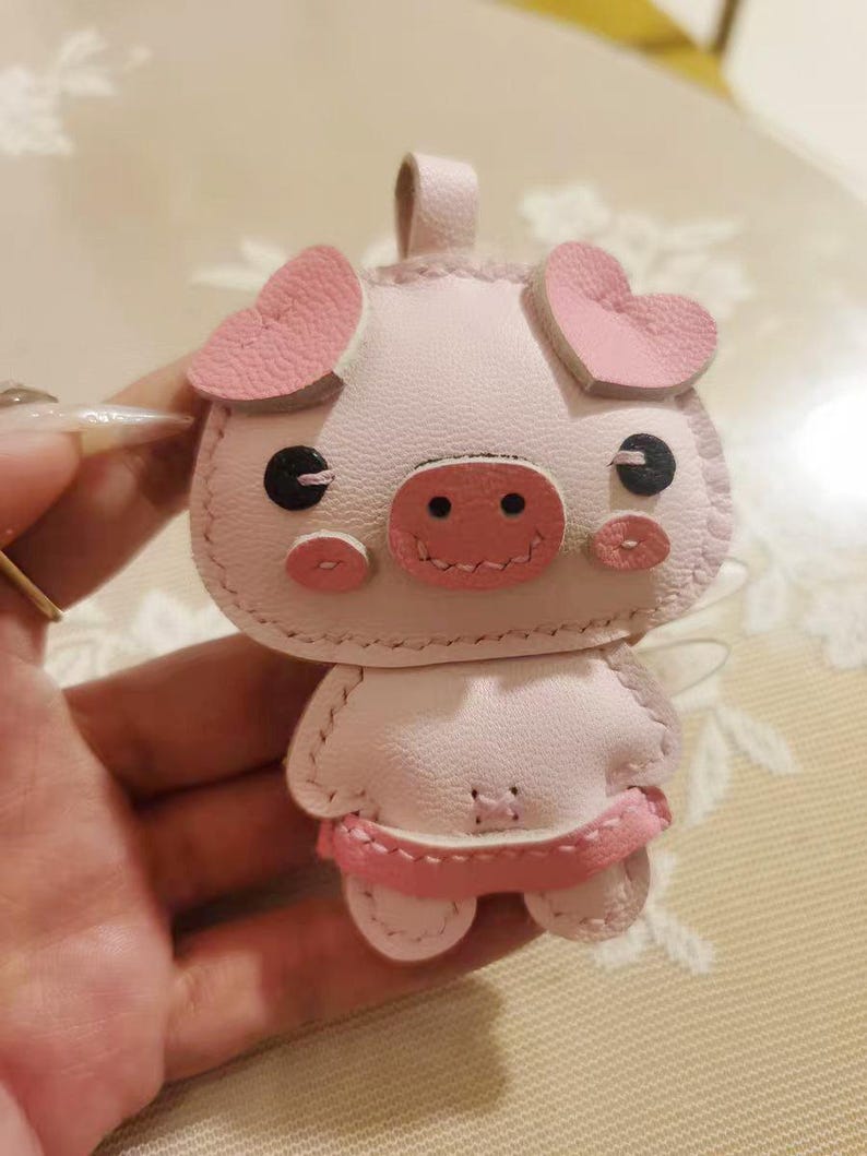 May include: A pink pig keychain made of leather with pink heart-shaped ears and a pink snout. The pig has black eyes and a stitched smile.
