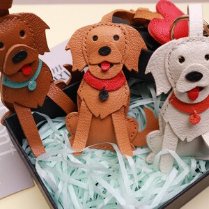 May include: Three leather dog keychains in shades of brown and off-white, each with a red or blue collar and a red tongue. The keychains are in a black box with shredded paper.