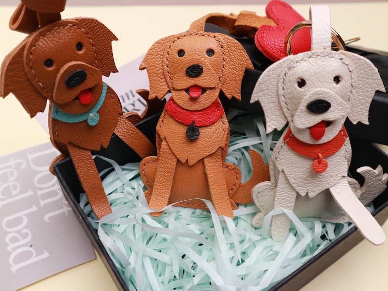 May include: Three leather dog keychains in shades of brown and off-white, each with a red or blue collar and a red tongue. The keychains are in a black box with shredded paper.