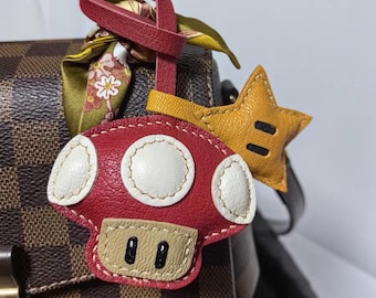May include: A red mushroom-shaped leather keychain with white accents and a tan base, paired with a yellow star. The keychain is attached to a red leather strap and a patterned silk scarf. The background is a brown checkered pattern.