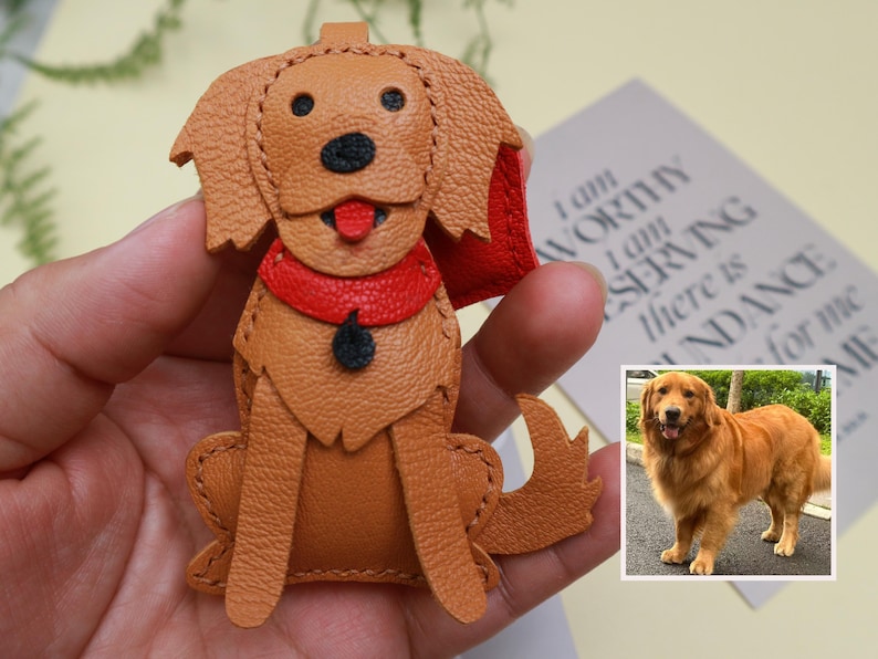 May include: A handcrafted leather dog keychain in a sitting position. The dog has a tan body, black nose, and red collar. The keychain is a charming accessory for dog lovers.