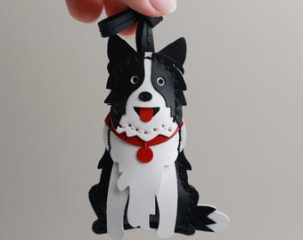 May include: A black and white leather border collie dog keychain with a red collar and a red button. The dog is sitting with its tongue sticking out.
