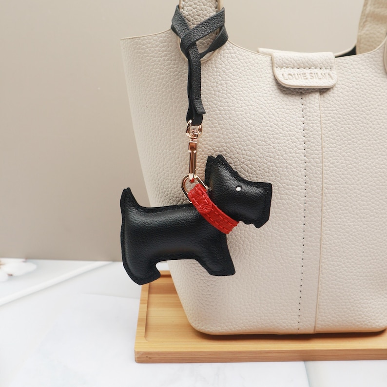 May include: A black leather dog-shaped keychain with a red collar and gold-tone hardware. The keychain is hanging from a light-colored handbag. The handbag has the text "LOUIE SILVA" embossed on it.