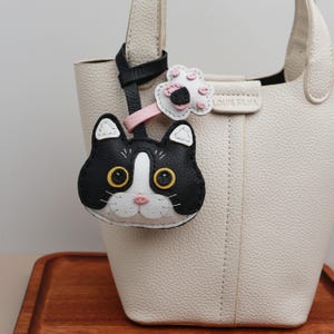 Può includere: Una borsa a mano color crema con un portachiavi a forma di gatto bianco e nero. Il portachiavi ha occhi gialli e un naso rosa. È attaccato anche un ciondolo a forma di fiore con dettagli rosa e neri. La borsa ha un manico superiore e il marchio "LOUIE SILVA" è visibile.