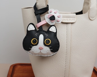 May include: A cream-colored handbag with a black and white cat-shaped keychain. The keychain has yellow eyes and a pink nose. A flower-shaped charm with pink and black accents is also attached. The bag has a top handle and the brand name "LOUIE SILVA" is visible.