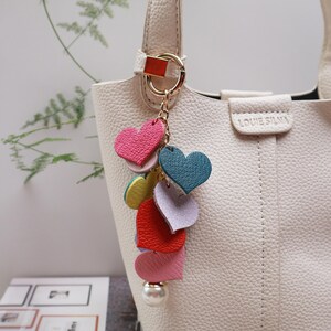 May include: A white handbag with a gold chain and a charm made of colorful leather hearts. The charm has a pearl at the bottom.