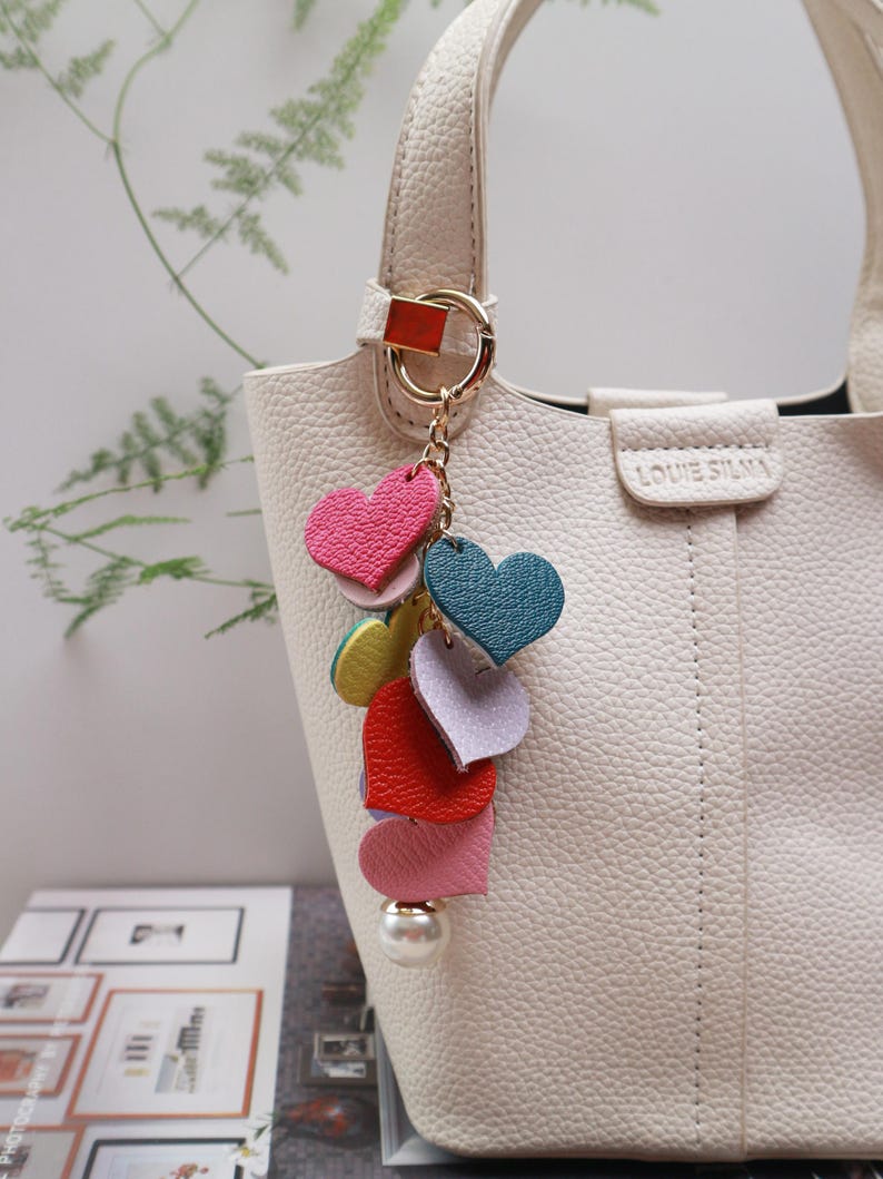 May include: A white handbag with a gold chain and a charm made of colorful leather hearts. The charm has a pearl at the bottom.