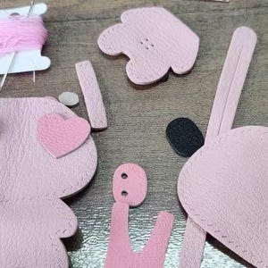 May include: Pink leather pieces for a DIY craft project to make a small stuffed animal. The pieces include a body, two hearts, two ears, a head, and a tail.