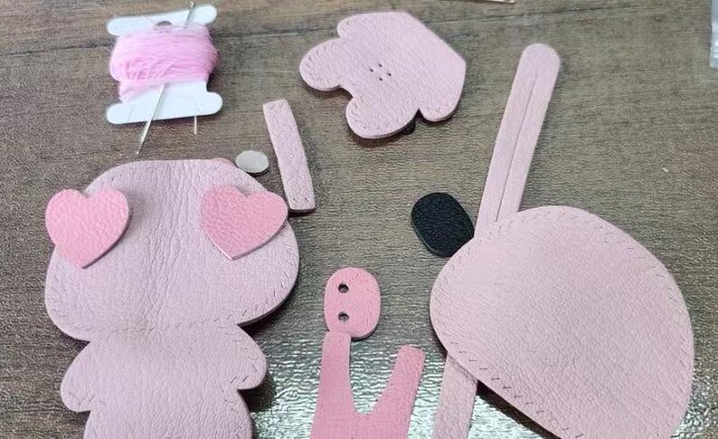 May include: Pink leather pieces for a DIY craft project to make a small stuffed animal. The pieces include a body, two hearts, two ears, a head, and a tail.