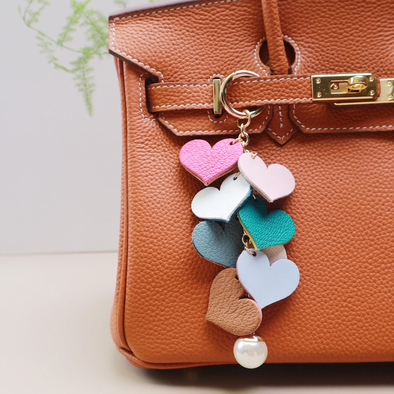 May include: A colorful leather heart charm with a pearl accent hangs from a gold key ring. The charm is attached to a brown leather handbag.