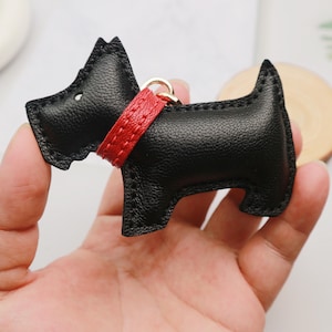 May include: Black leather dog-shaped keychain with a red collar and gold-colored ring. The keychain is a stylized representation of a Scottish Terrier. The dog has a textured surface and is held in a hand.