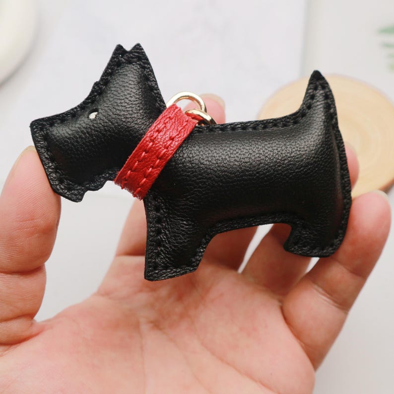 May include: Black leather dog-shaped keychain with a red collar and gold-colored ring. The keychain is a stylized representation of a Scottish Terrier. The dog has a textured surface and is held in a hand.