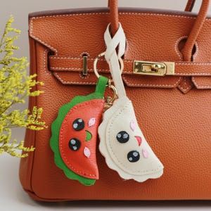 May include: A brown leather handbag with a gold clasp and a key chain with two charms. One charm is a red and green watermelon and the other is a white dumpling.