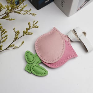 May include: A collection of leather accessories in the shape of leaves and petals. The main pieces are pink, with a white leather strap and green leaf accents. The items are stitched with a contrasting thread, suggesting a handmade craft.