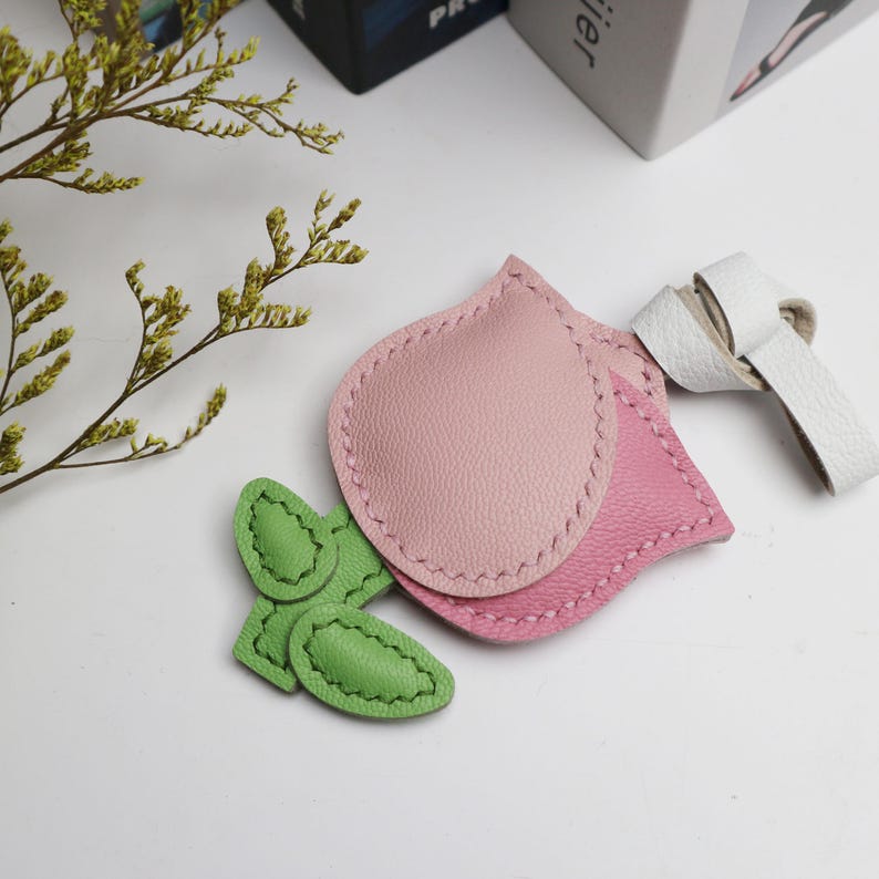 May include: A collection of leather accessories in the shape of leaves and petals. The main pieces are pink, with a white leather strap and green leaf accents. The items are stitched with a contrasting thread, suggesting a handmade craft.