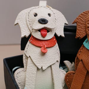 May include: A white leather dog-shaped keychain with a red collar and tongue. The dog has black eyes and a black nose. The keychain is sitting in a black box with light blue tissue paper.