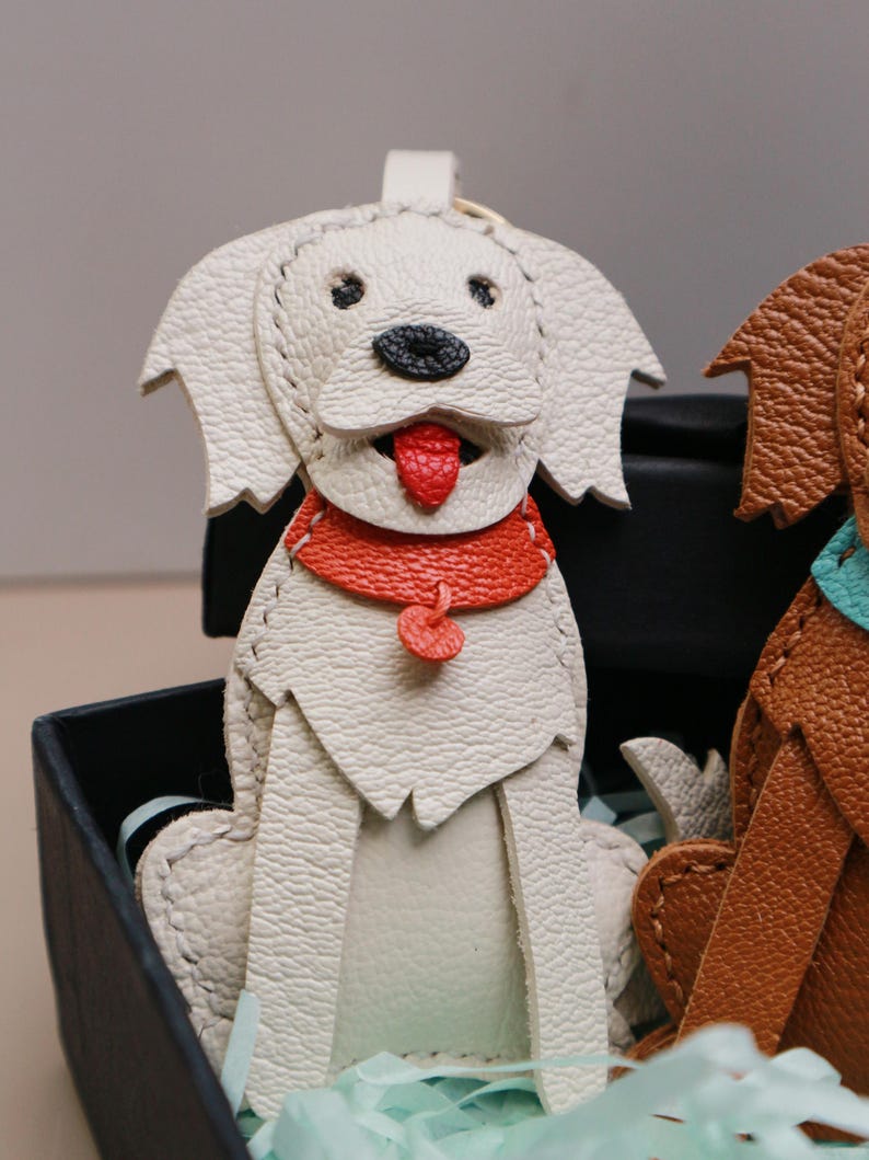 May include: A white leather dog-shaped keychain with a red collar and tongue. The dog has black eyes and a black nose. The keychain is sitting in a black box with light blue tissue paper.