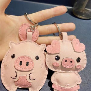 May include: Two pink pig-shaped leather keychains with black eyes and stitched details. One pig is standing with heart-shaped ears and the other is sitting with round ears.