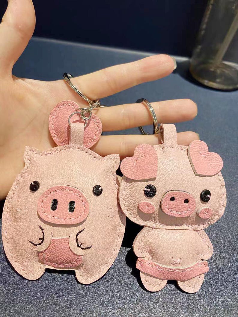 May include: Two pink pig-shaped leather keychains with black eyes and stitched details. One pig is standing with heart-shaped ears and the other is sitting with round ears.