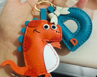 May include: A leather keychain featuring a bright orange dinosaur and a blue letter D. The dinosaur has white accents and black eyes. The keychain is attached to a gold ring.