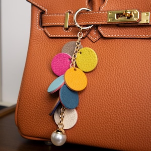 May include: A colorful leather keychain with a pearl charm. The keychain features a series of round leather pieces in pink, yellow, blue, and green, attached to a gold chain.