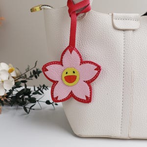 Puede incluir: Un bolso tote de cuero blanco con un dije de flor rosa. La flor tiene un contorno rojo y una carita sonriente amarilla en el centro.