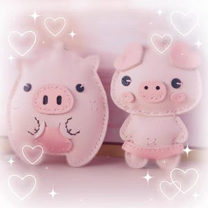 以下が含まれることがあります： ピンクの豚の形をした革製のキーホルダーが2つ。1つの豚は立っていて、もう1つの豚は座っています。両方の豚は黒い目とピンクの鼻をしています。