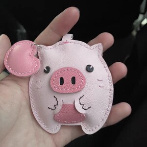 May include: A pink leather pig keychain with a heart-shaped charm. The pig has black eyes and a pink snout.