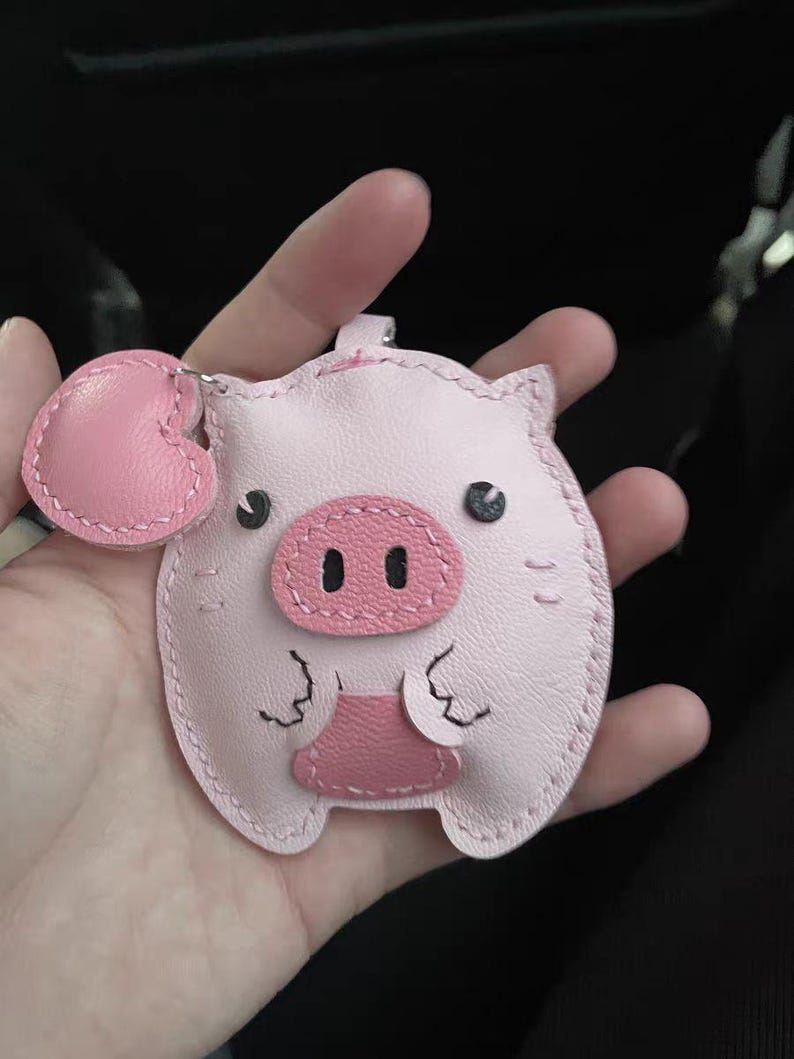May include: A pink leather pig keychain with a heart-shaped charm. The pig has black eyes and a pink snout.
