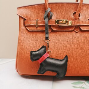 May include: A black leather dog-shaped keychain with a red collar hangs from a gold clasp. The keychain is attached to a brown leather handbag with gold hardware. The handbag has a textured surface and a top handle.