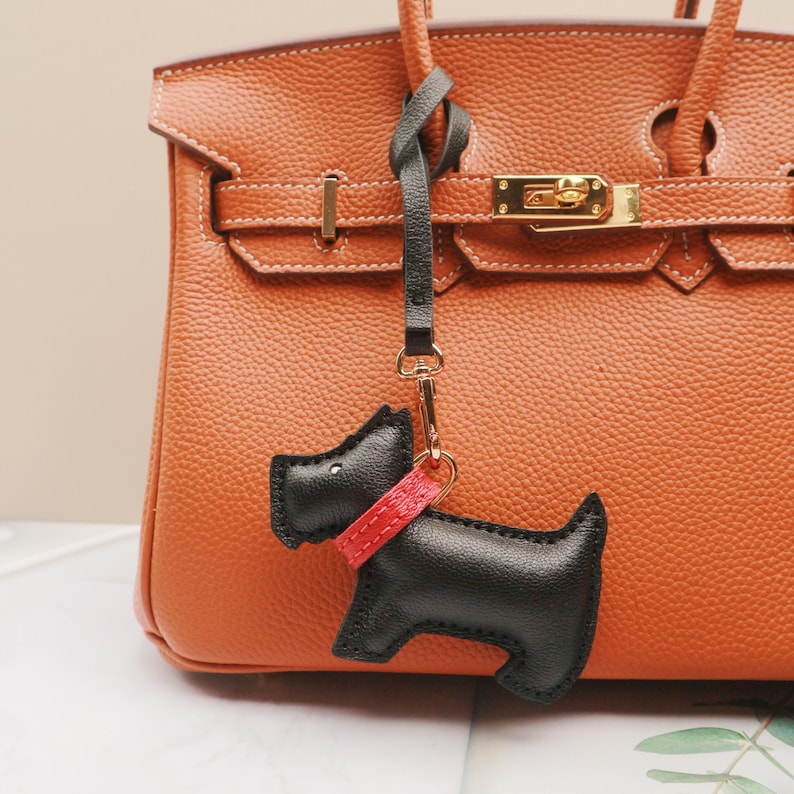 May include: A black leather dog-shaped keychain with a red collar hangs from a gold clasp. The keychain is attached to a brown leather handbag with gold hardware. The handbag has a textured surface and a top handle.