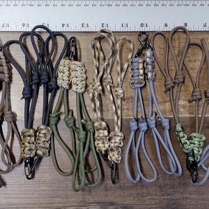 May include: A collection of 16 colorful paracord keychains with a variety of patterns and colors, including camouflage, pink, purple, and teal. Each keychain has a metal clip.