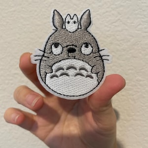 May include: Embroidered patch featuring a gray Totoro character with a smaller Totoro on its head. The patch has a white border and detailed stitching, showcasing the character's eyes, whiskers, and belly. A cute and whimsical design.