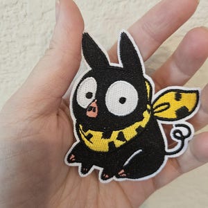 May include: Embroidered patch featuring a black cartoon pig-like creature with large white eyes, a pink snout, and a curly tail. The creature wears a yellow collar and has yellow wings. The patch is designed with black and yellow thread.
