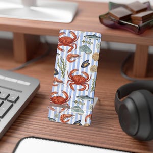 Può includere: Un supporto per telefono bianco con un motivo a righe blu e bianche con illustrazioni di granchi, gamberi, pesci, conchiglie e alghe. Il supporto è progettato per contenere un telefono.