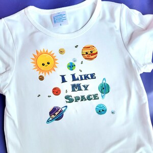 May include: White t-shirt with a graphic of a smiling sun and planets with the text "I Like My Space".