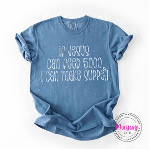 May include: A blue t-shirt with the text "if Jesus can feed 5000, I can make supper" in white script. The shirt has short sleeves and a crew neck. A logo is visible in the bottom right corner.
