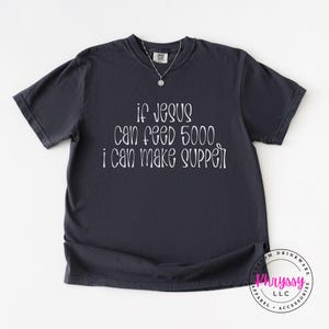 May include: Dark gray t-shirt with the text "If Jesus can feed 5000, I can make supper" in white script. The shirt has a crew neck and short sleeves. A silver necklace with a small pendant is visible.