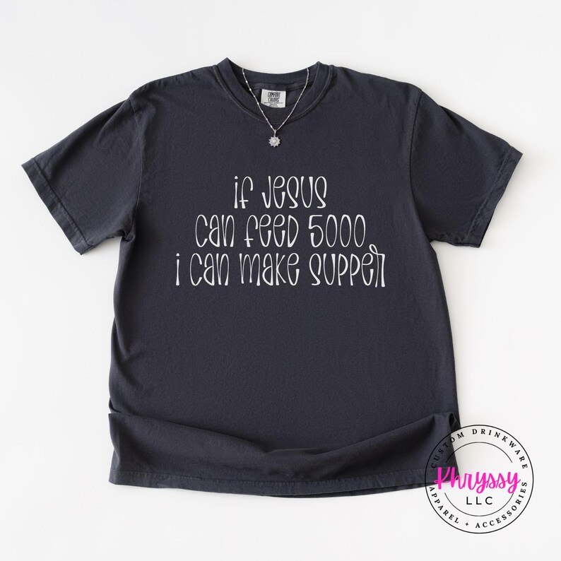 May include: Dark gray t-shirt with the text "If Jesus can feed 5000, I can make supper" in white script. The shirt has a crew neck and short sleeves. A silver necklace with a small pendant is visible.