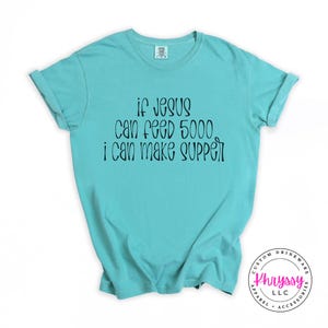 May include: A turquoise t-shirt with the text "if Jesus can feed 5000 i can make supper" in black script. The shirt has short sleeves and a crew neck. The bottom right corner has a circular logo with text.
