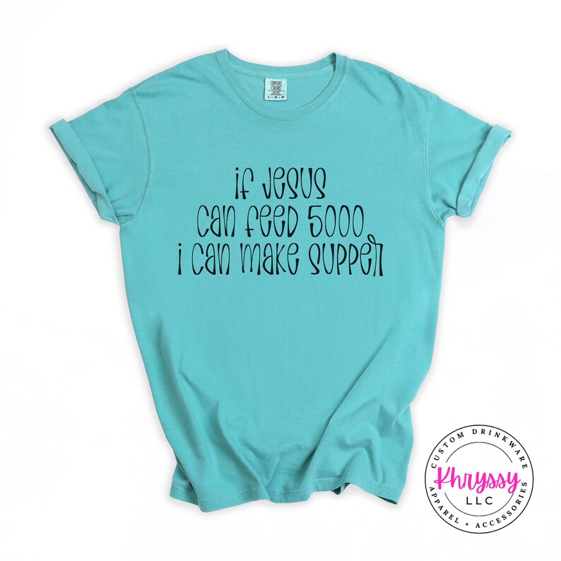May include: A turquoise t-shirt with the text "if Jesus can feed 5000 i can make supper" in black script. The shirt has short sleeves and a crew neck. The bottom right corner has a circular logo with text.