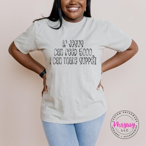 May include: A light beige t-shirt with the text "if Jesus can feed 5000, I can make supper" in black script. The shirt is a crew neck style and is made of a soft material.