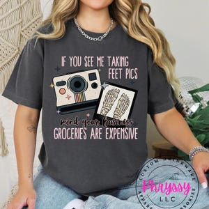 May include: A dark gray t-shirt with a graphic of a vintage camera and a skeleton foot. The text on the shirt reads "If you see me taking feet pics mind your business groceries are expensive".