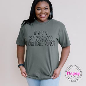 May include: An olive green t-shirt with the text "if Jesus can feed 5000, I can make supper" in black script. The shirt is worn by a person. A logo for "Khryssy LLC" is visible on the right side of the image.