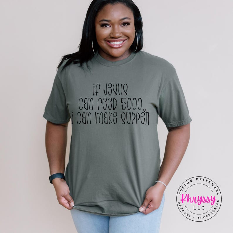 May include: An olive green t-shirt with the text "if Jesus can feed 5000, I can make supper" in black script. The shirt is worn by a person. A logo for "Khryssy LLC" is visible on the right side of the image.